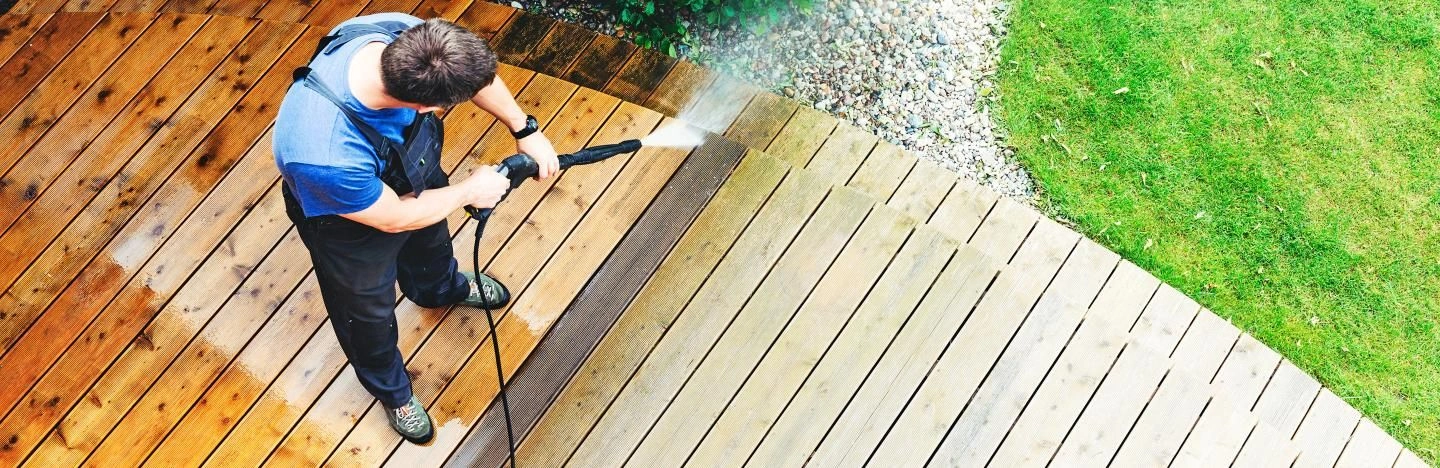 A landscape image of a man using a jet wash to clean their decking in their garden, making sure to use it 2-4 feet away from the actual decking to not damage it. 