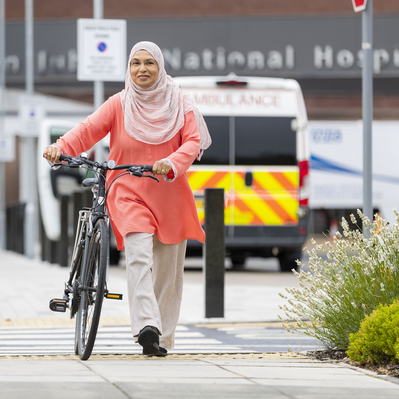 a woman wearing a hijab is carrying a bike with a parked ambulance in the background