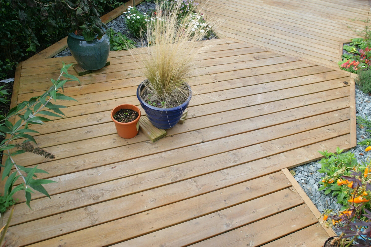 Decking in a garden with plant pots, shrubbery and grass and gravel surrounding it.
