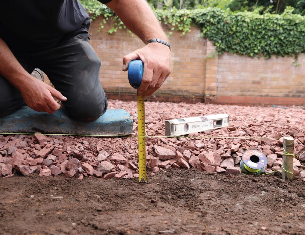 A person using tape measures and pegs to figure out the fall of the patio, which is used to allow water to drift off of the patio. 