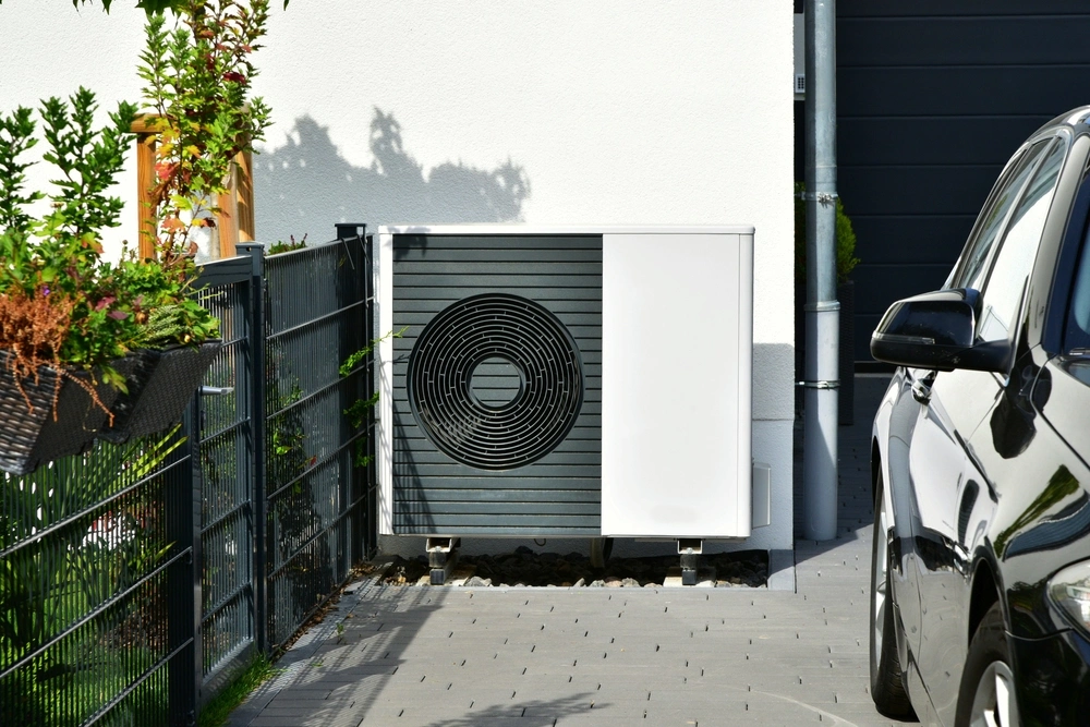 Air Source Heat Pump located outside someones home and against a wall with a car being shown on  the right and a fence on the left.