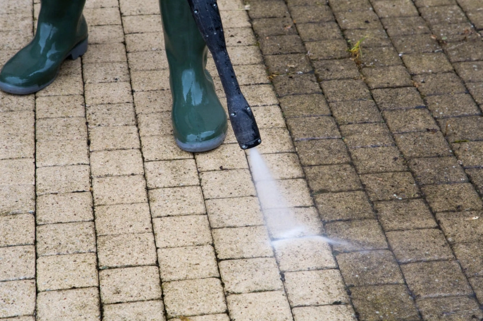 An image of a person with green boots using a jet wash to clean the block paving beneath them. 