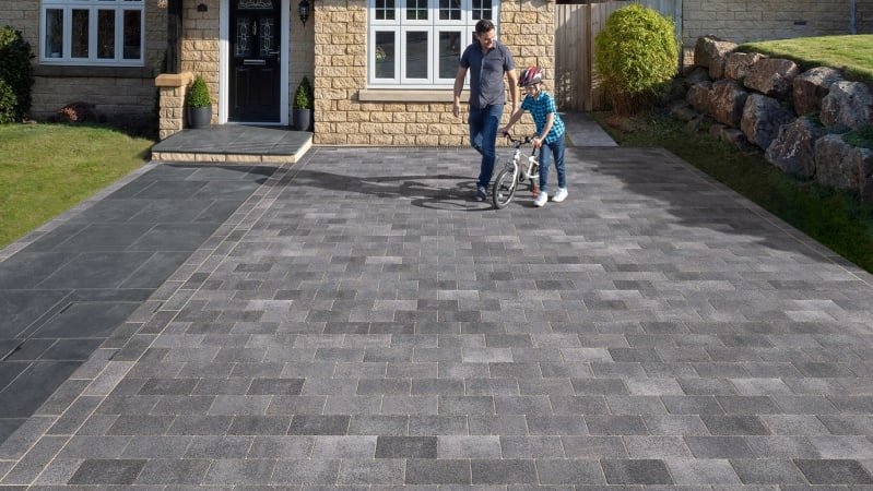 An image of a man and their child riding a bike on the Marshalls Nova Coarse block paving, which has been used as a driveway for their house behind them. 