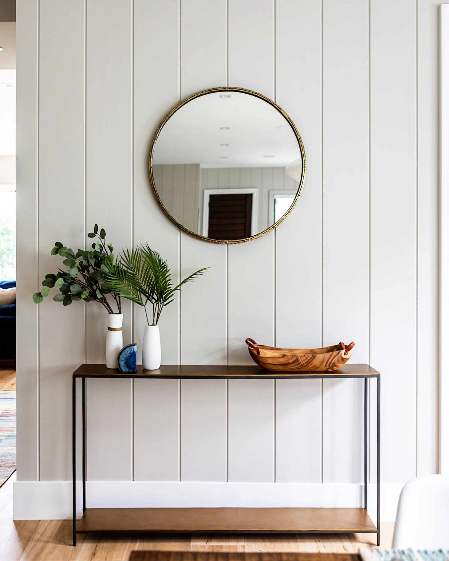Bright white shiplap wall panelling used in a hallway and decorated with a table, plant and gold-coloured mirror.