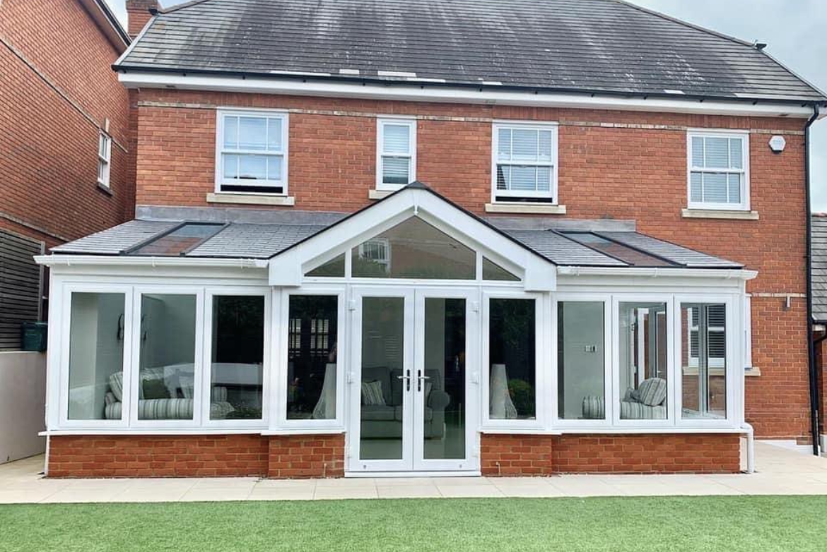 Showing a detached property from the back garden looking into the conservatory extension. Slanted extension roof and double doors into the home.