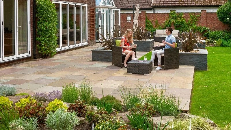 A landscape picture of a couple sat on garden furniture on top of a patio in the garden. 