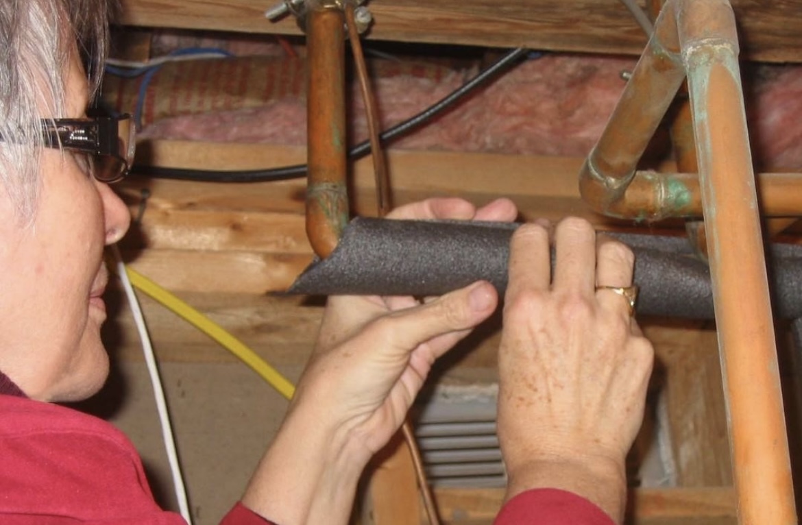 An image showing a woman installing pipe insulation in the loft. Close up of copper pipes with grey pipe insulation wrapped around.