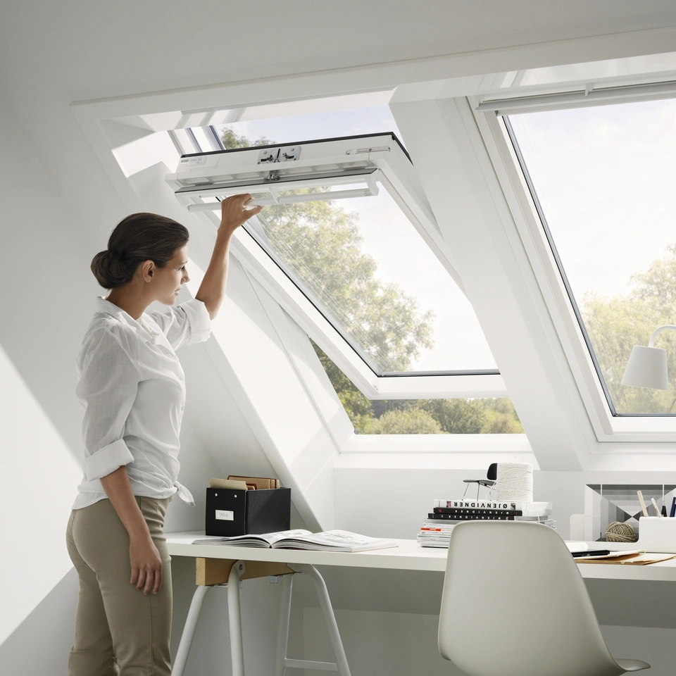A woman opening one of Velux's roof windows to let more light in. Plain white walls and white window.
