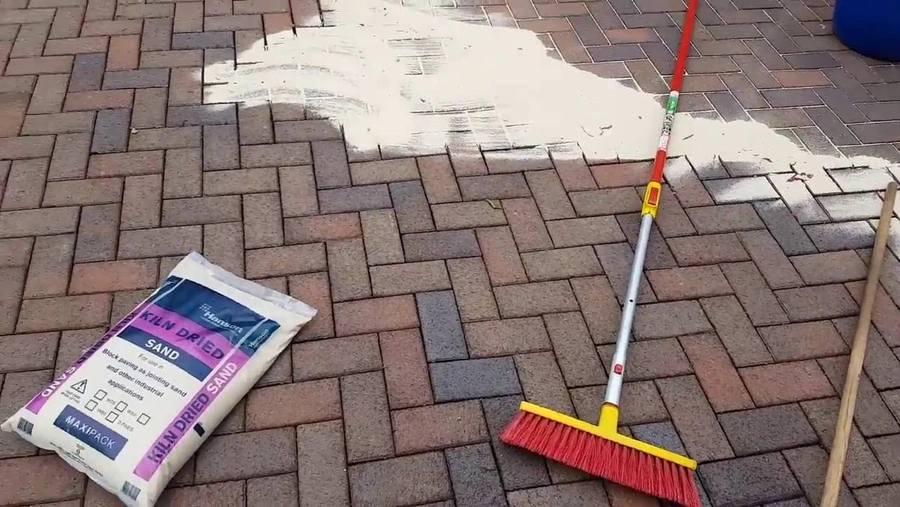 An image of kiln-dried sand and a bristle brush being used to maintain and clean a block paving driveway. 