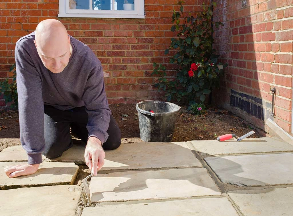 A person using a bucket and spade to apply fresh mortar in between the cracks of paving slabs for a patio to grout them.
